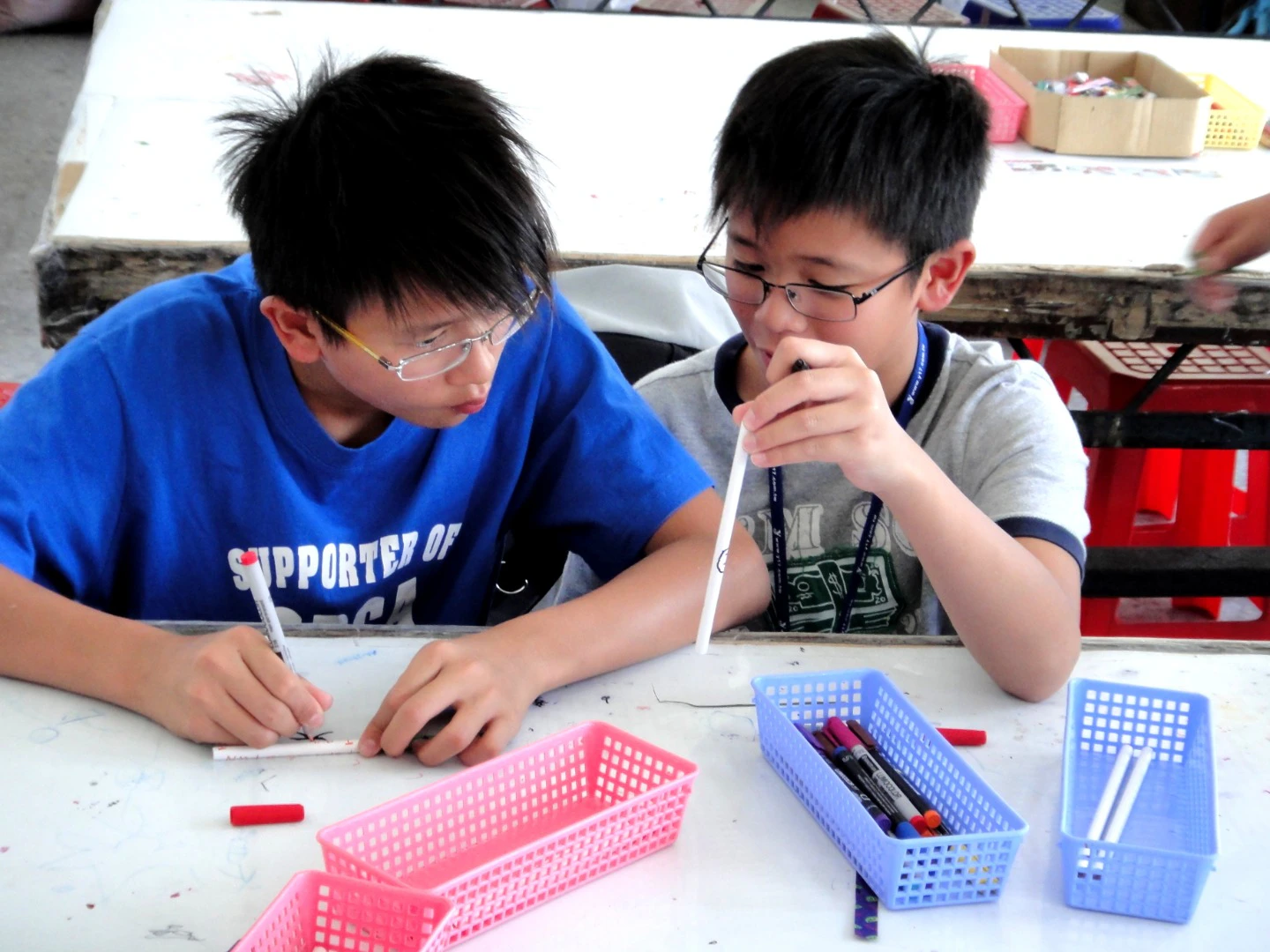 Two children sitting at a table working on a project with markers and colorful baskets containing supplies.