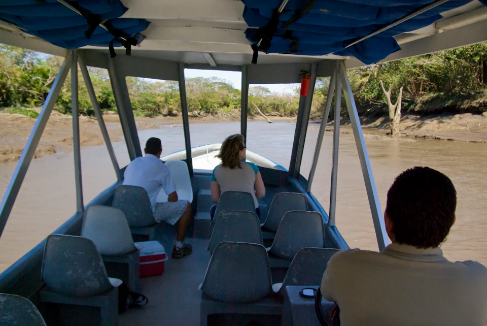 Three people sit on a small boat navigating a narrow, tree-lined river under a covered roof, heading away from the viewer.