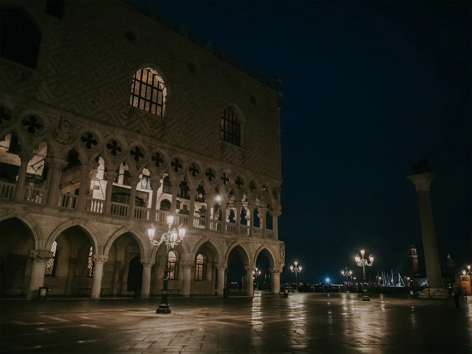 Illuminated historic building at night with arched windows and columns; street lamps light up the empty cobblestone square.
