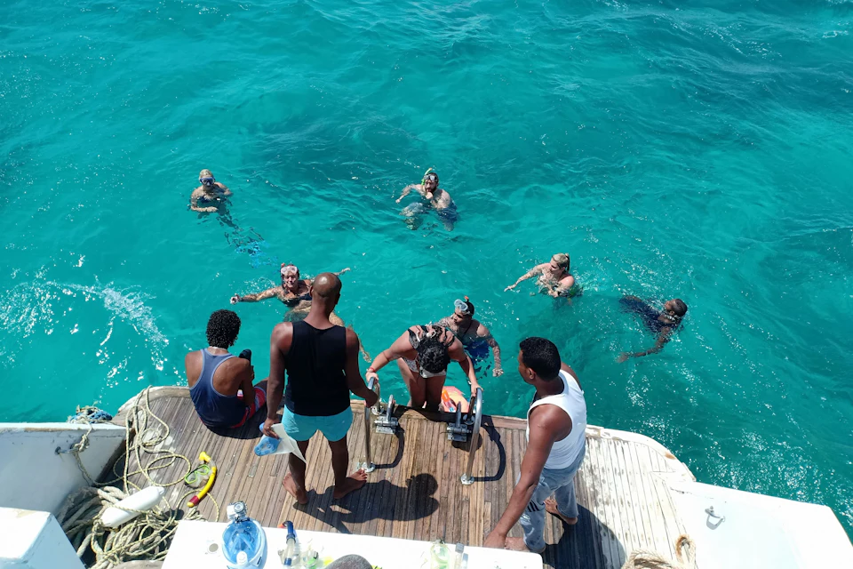 People enjoying a swim in clear blue water next to a boat. Some are onboard, while others are in the water near the boat.