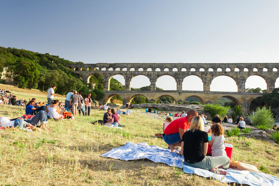 People picnicking on a grassy field near an ancient Roman aqueduct with multiple arches on a clear day.