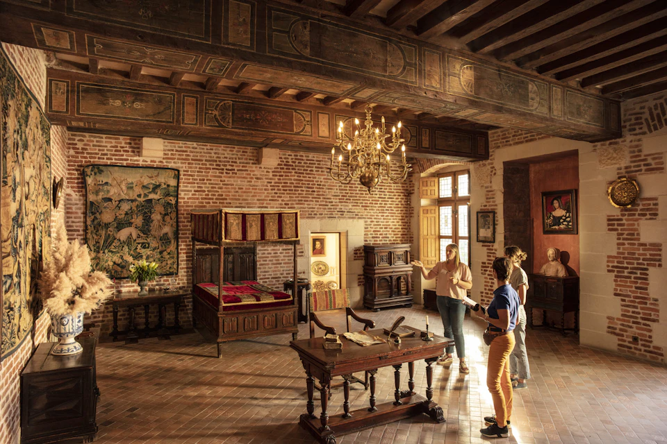 A historical room with ornate furniture, a chandelier, and three people observing and discussing the decor.
