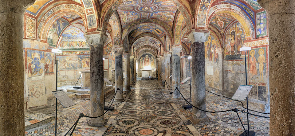 A historic chapel with intricate mosaic floors, stone columns, and richly detailed frescoed ceilings and walls.
