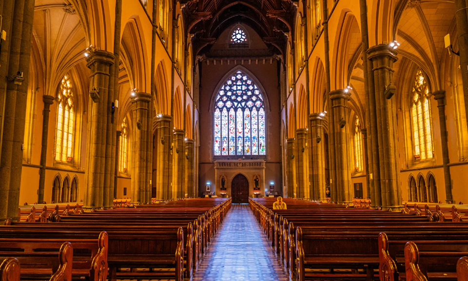 Interior of a cathedral with a stained glass window, rows of wooden pews, high arches, and a single person sitting near the front.