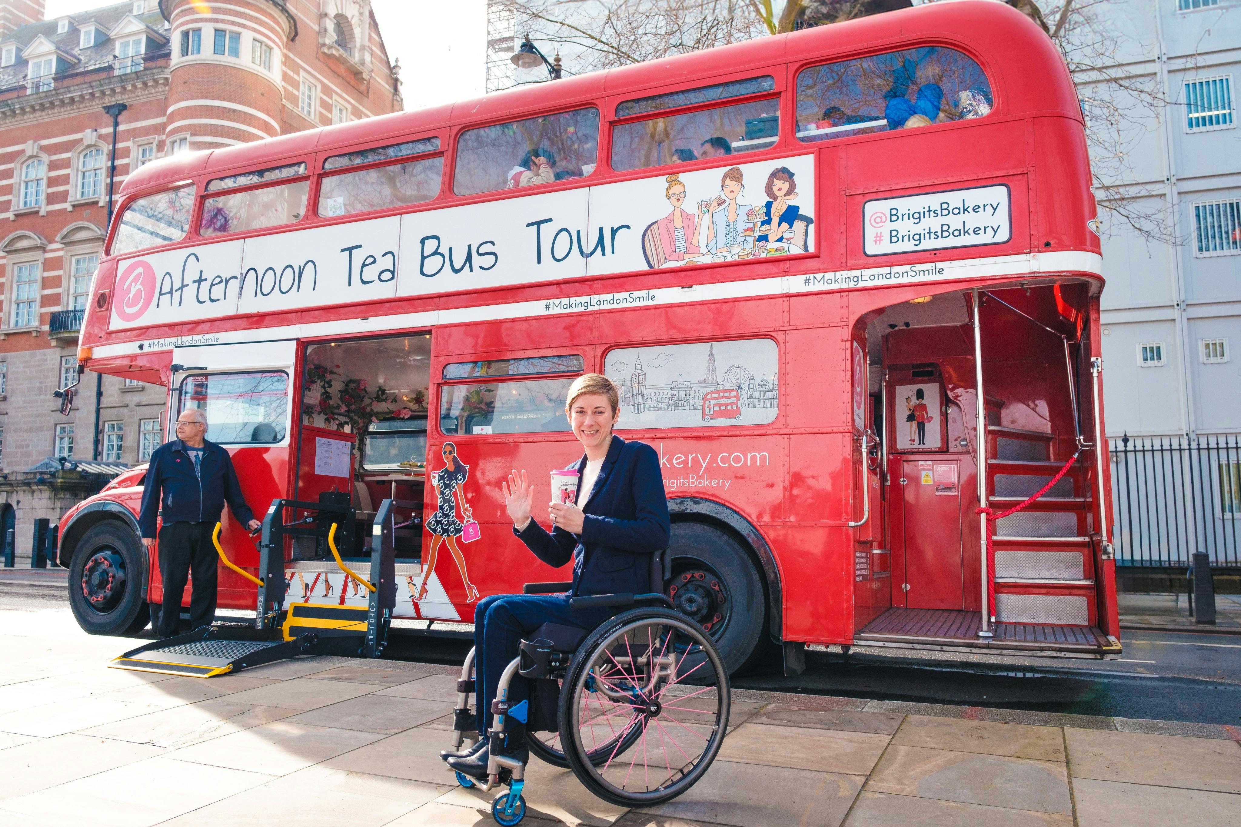 Osoba na wózku inwalidzkim uśmiechająca się i machająca przed czerwonym piętrowym autobusem z napisem „Wycieczka autobusem po popołudniowej herbacie”.