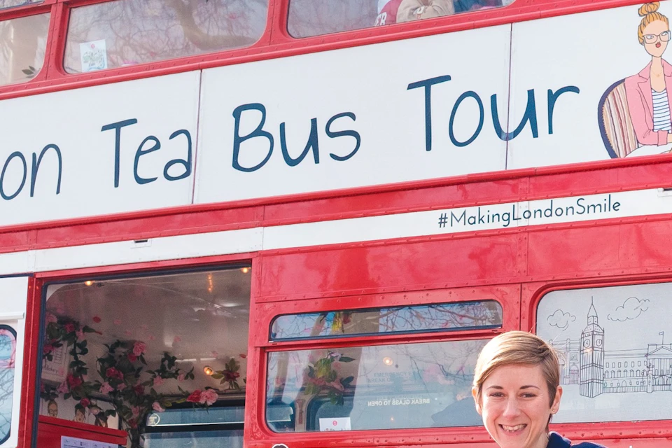 A person in a wheelchair smiling and waving in front of a red double-decker bus labeled 'Afternoon Tea Bus Tour.'