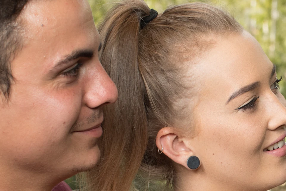 A man and woman smiling together outdoors, with foliage and a tree trunk nearby.