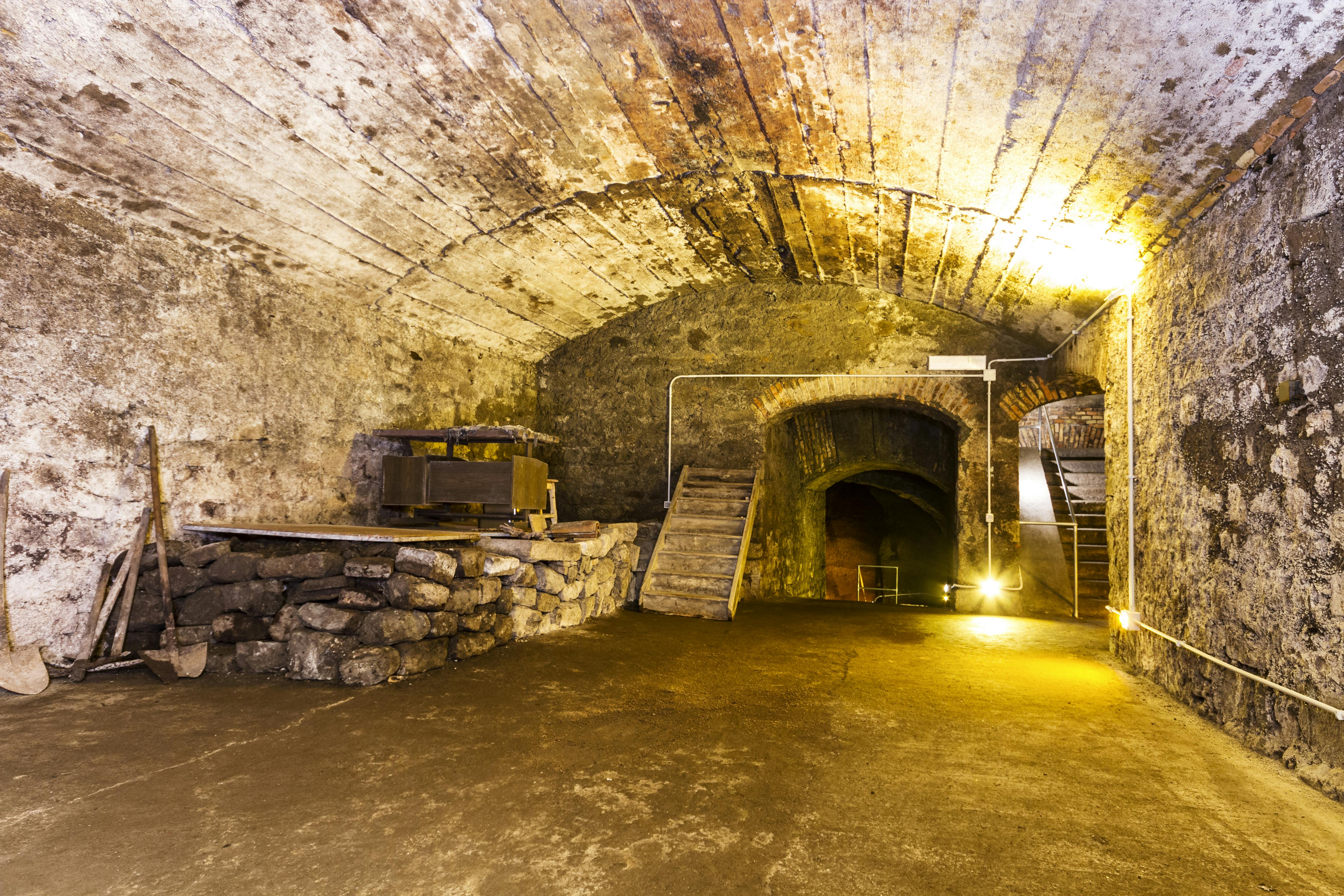 En gammel stenkælder med buede lofter, trapper, en hævet platform med maskiner og svag belysning.