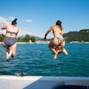 Two women in swimsuits jump off a boat into a lake, with hills and distant buildings under a clear blue sky in the background.