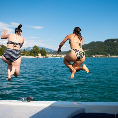 Two women in swimsuits jump off a boat into a lake, with hills and distant buildings under a clear blue sky in the background.