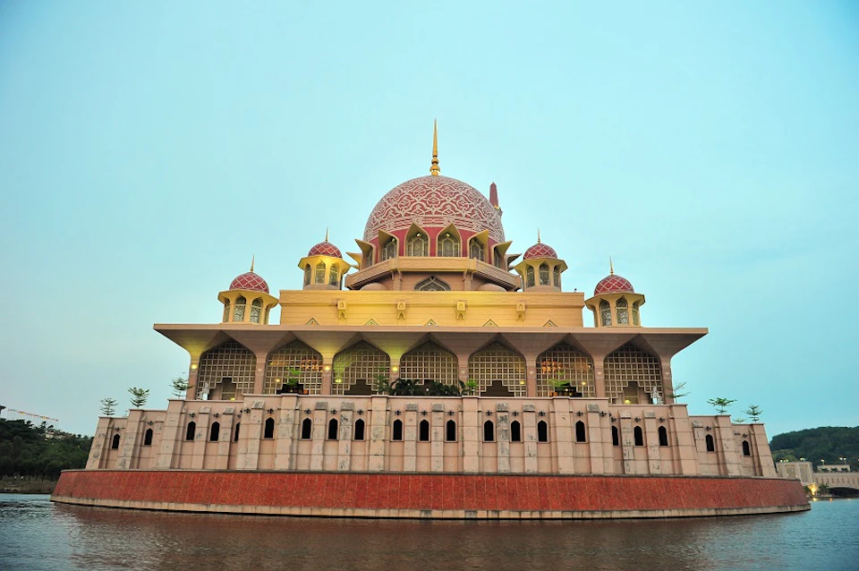 A mosque with a large pink dome and four smaller domes, featuring arched windows, viewed from across a body of water.