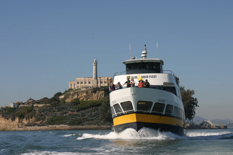 A ferry labeled "Blue & Gold Fleet" approaches with passengers on board, Alcatraz Island and its lighthouse are in the background.