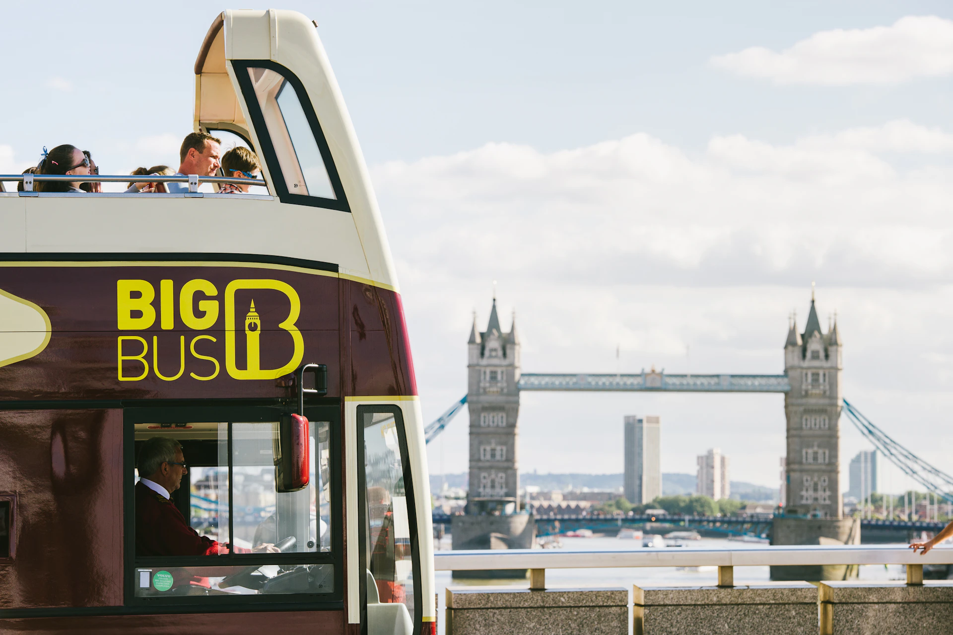 Double-decker tour bus with "BIG BUS" logo, people on upper deck, Tower Bridge in the background under a partly cloudy sky.