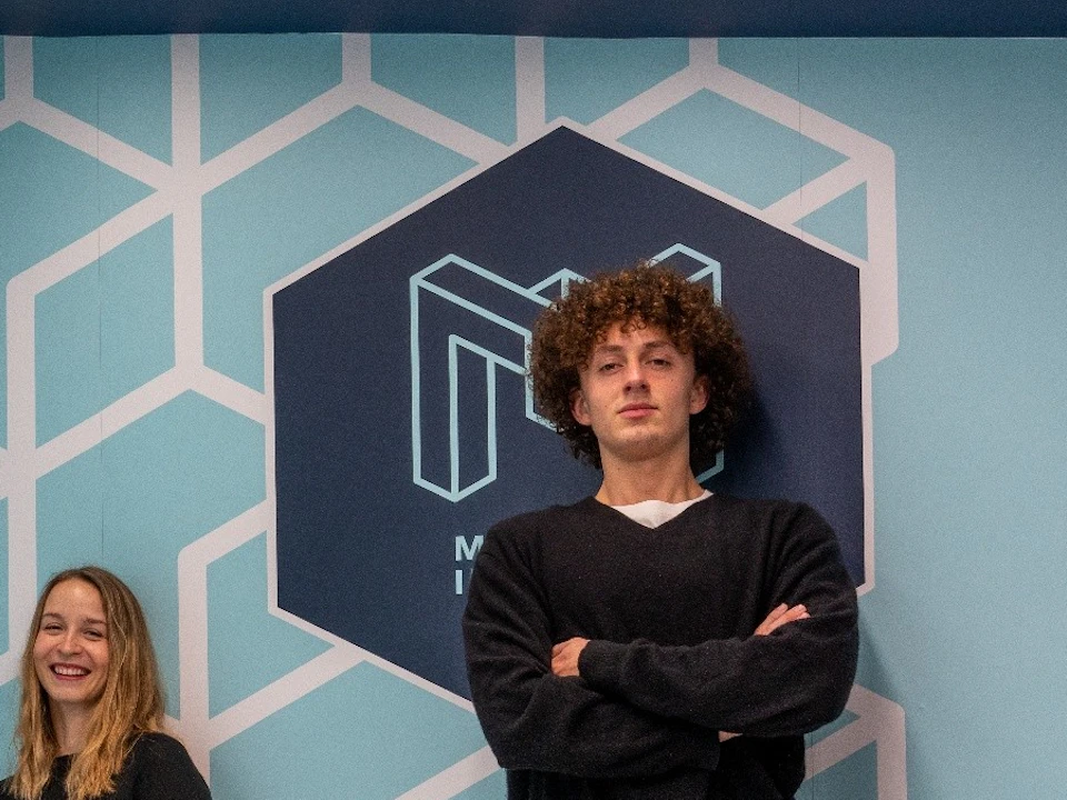 Four people in a room with a geometric patterned wall, posing in a way that creates a perspective optical illusion.