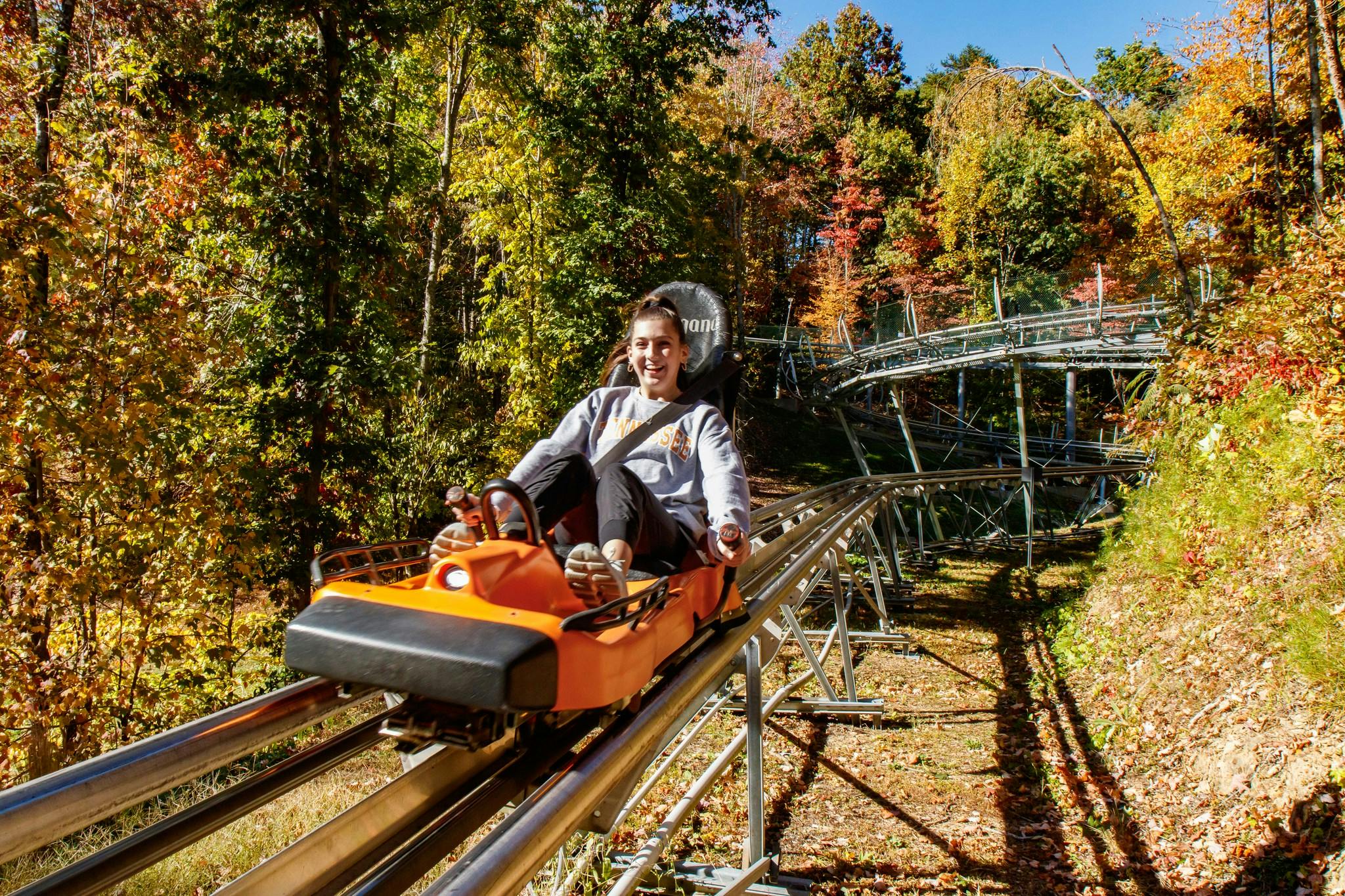 Person smiling while riding a single-seat, orange roller coaster cart through a wooded area with colorful autumn foliage.
