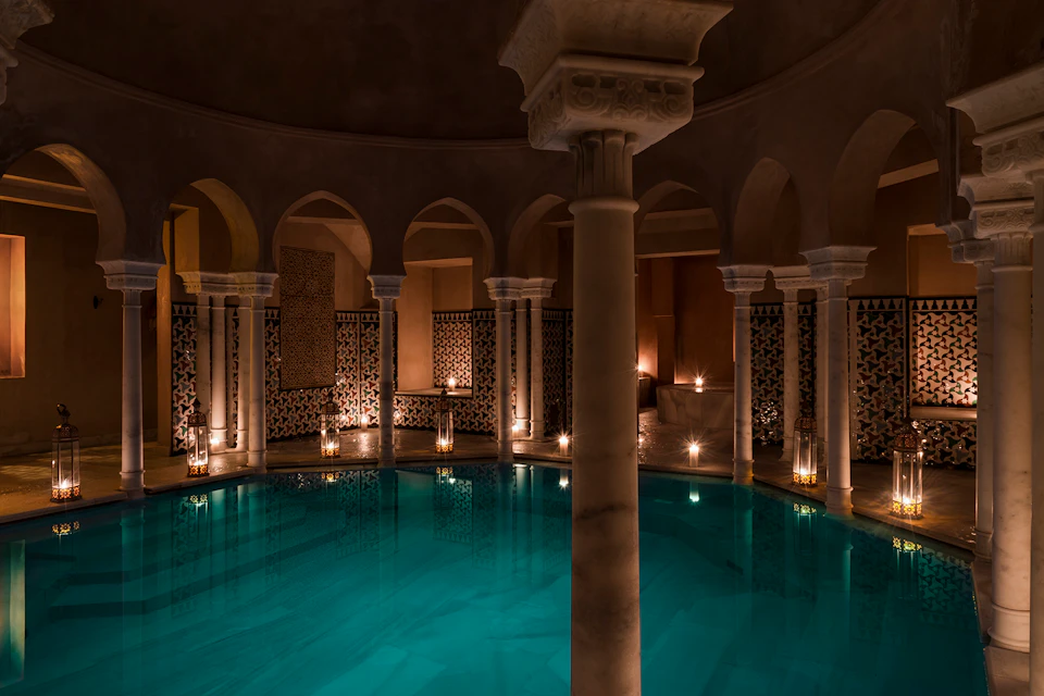 Dimly lit indoor pool with arched doorways and columns, surrounded by patterned tiles and illuminated by candles.