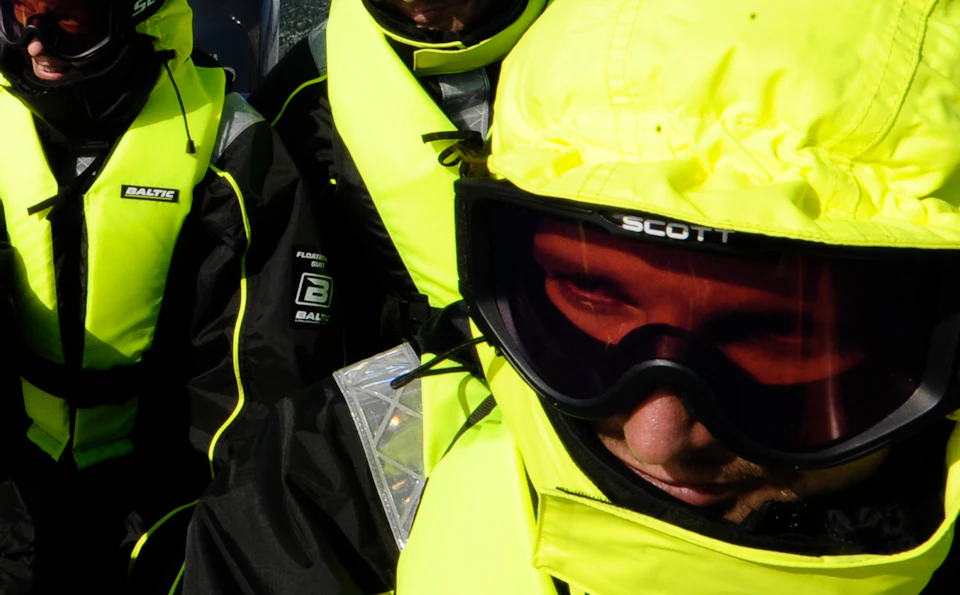 Group of people in bright yellow safety gear and goggles on a speeding boat in open water, with a clear blue sky.