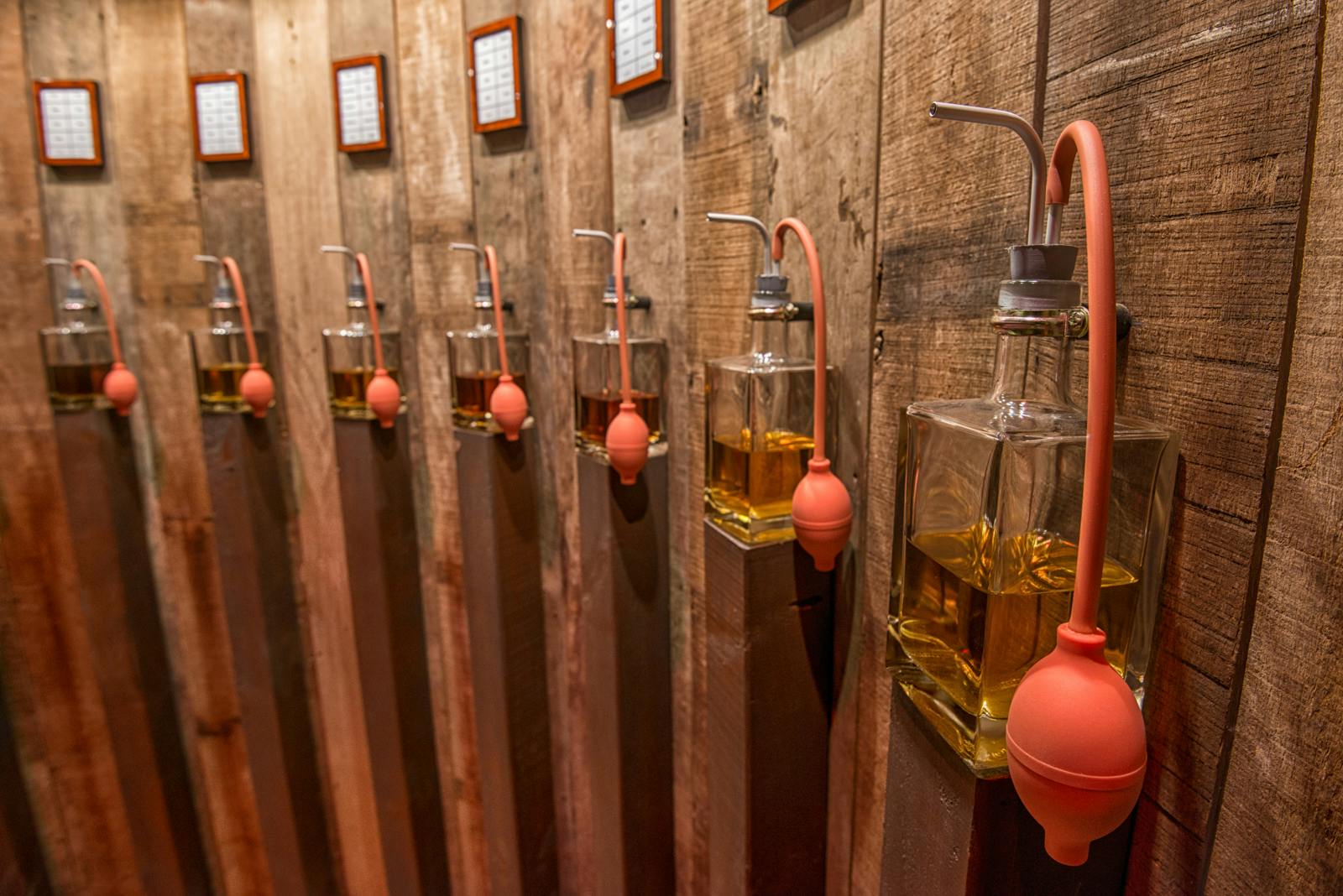 Řady skleněných zásobníků s oranžovými žárovkami, z nichž každá je naplněna jantarovou kapalinou, namontované na rustikálních dřevěných stěnových panelech.