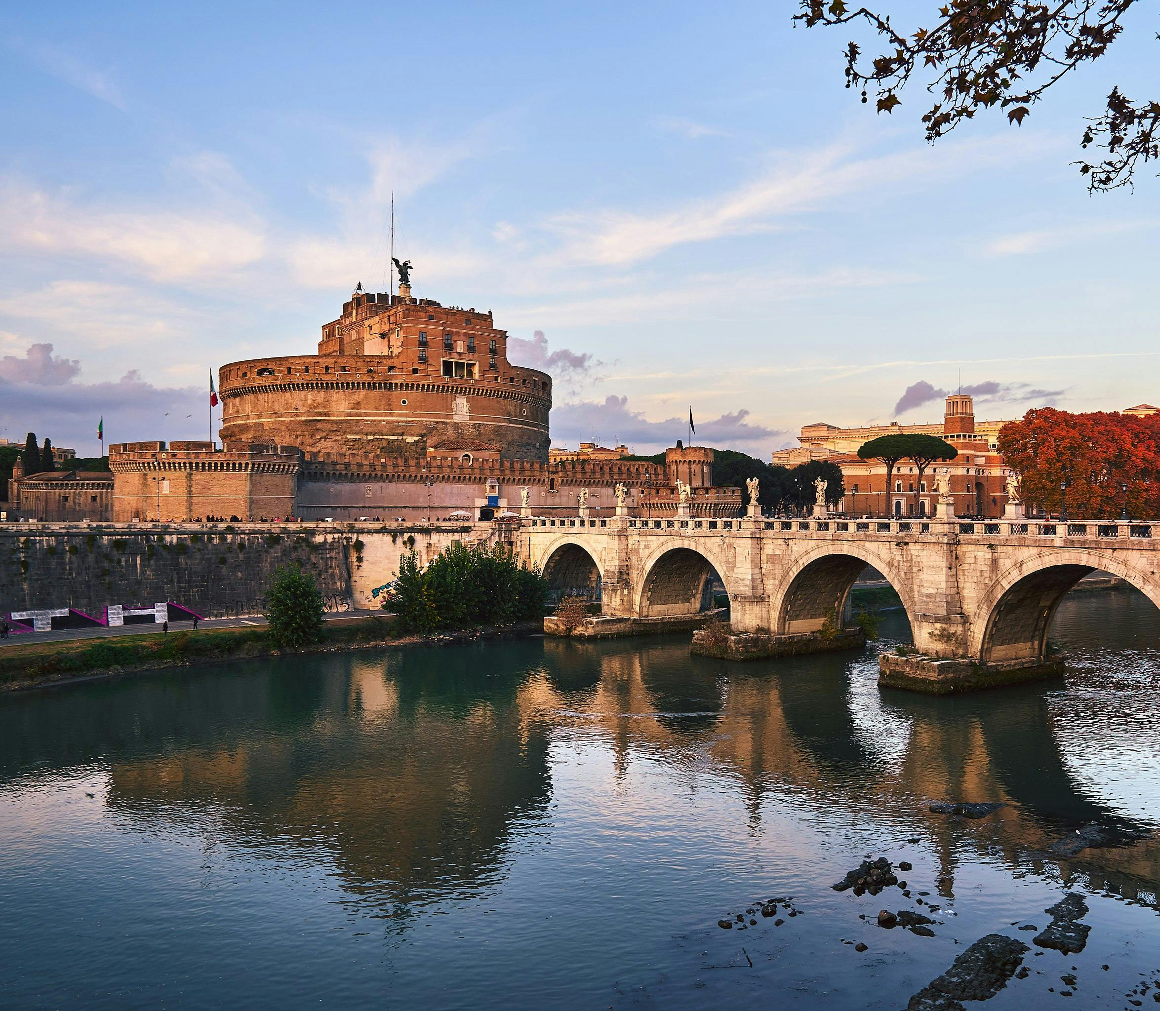 A historic stone bridge over a calm river with a large, ancient fortress in the background under a pastel-colored sunset sky.