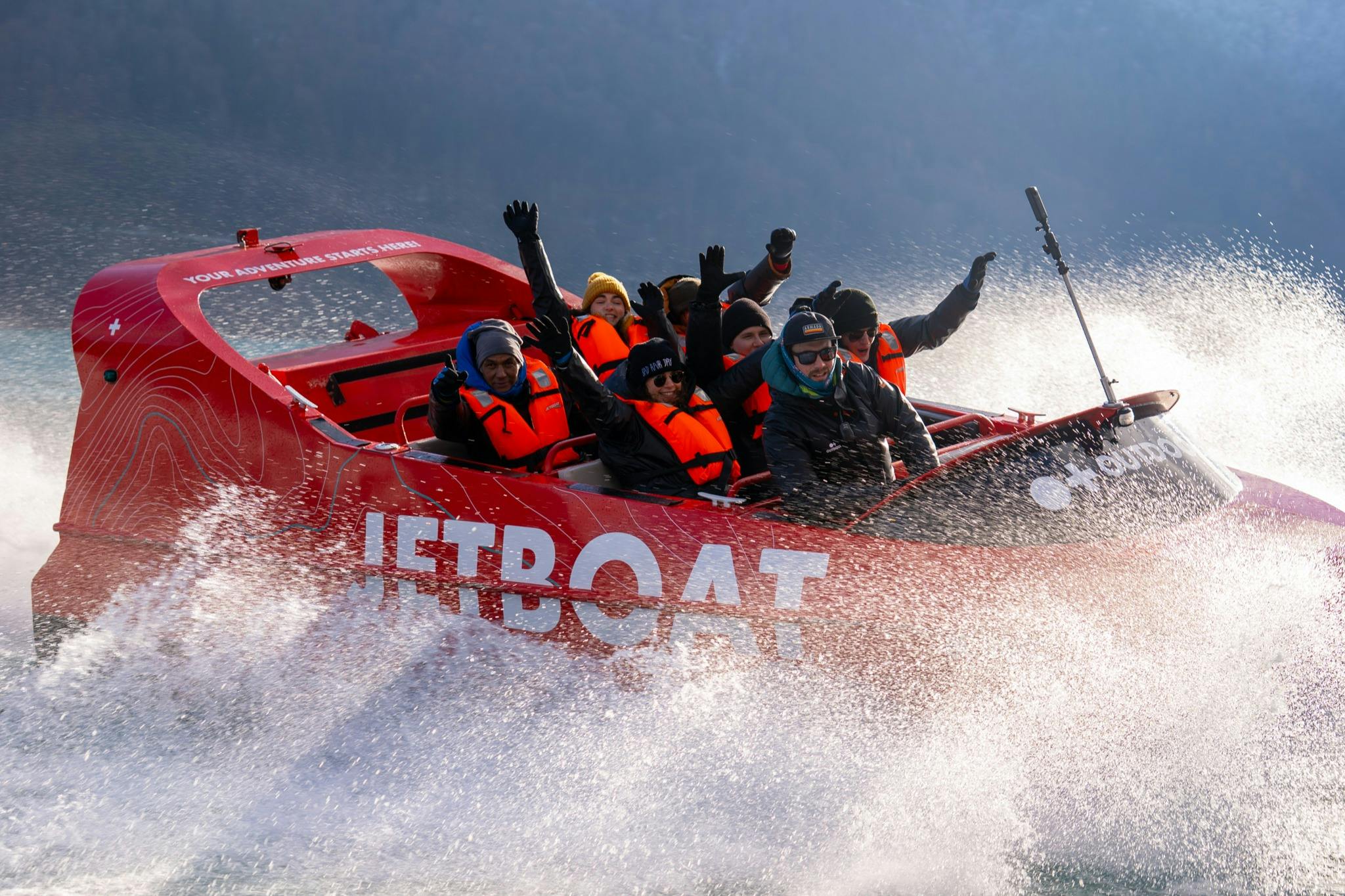 A group of people wearing life jackets enjoying a ride on a red jet boat, splashing through water at high speed.