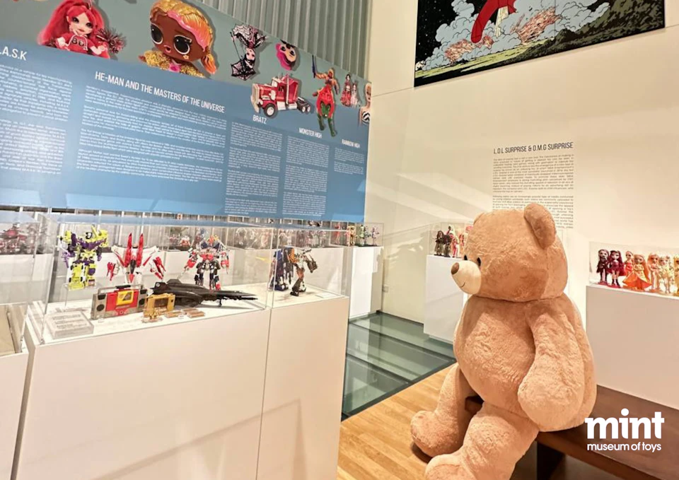 A museum exhibit displaying various action figures and toys, with an oversized teddy bear seated in the foreground.