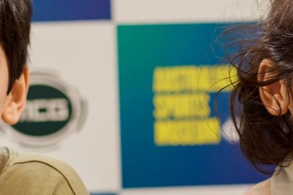 Two children sitting at a table with microphones, smiling, in front of a backdrop featuring the Australian Sports Museum logo.
