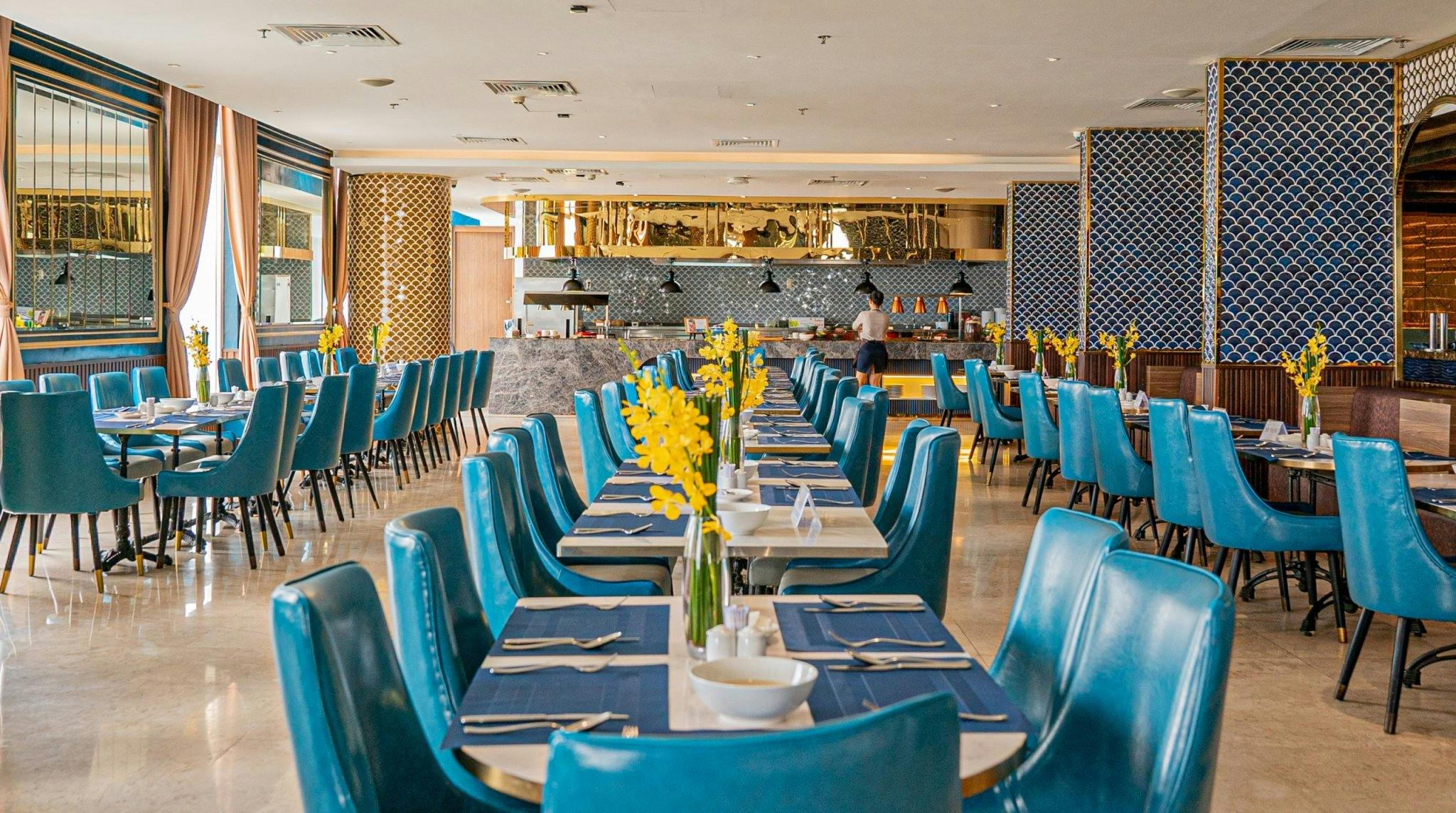 A restaurant dining area with blue chairs, tables set with plates and utensils, and yellow flowers as centerpieces.