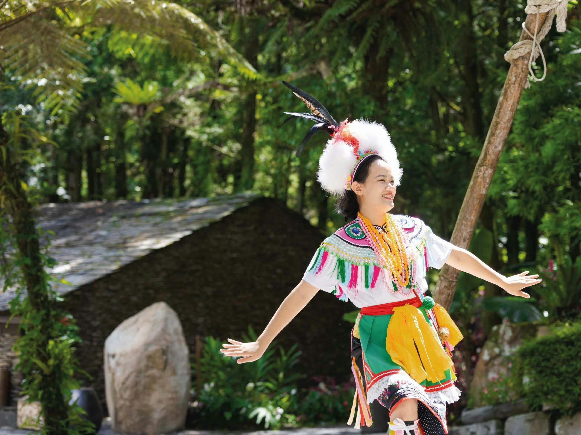 A person in colorful traditional attire dances outdoors in a lush green setting with trees and a wooden structure.