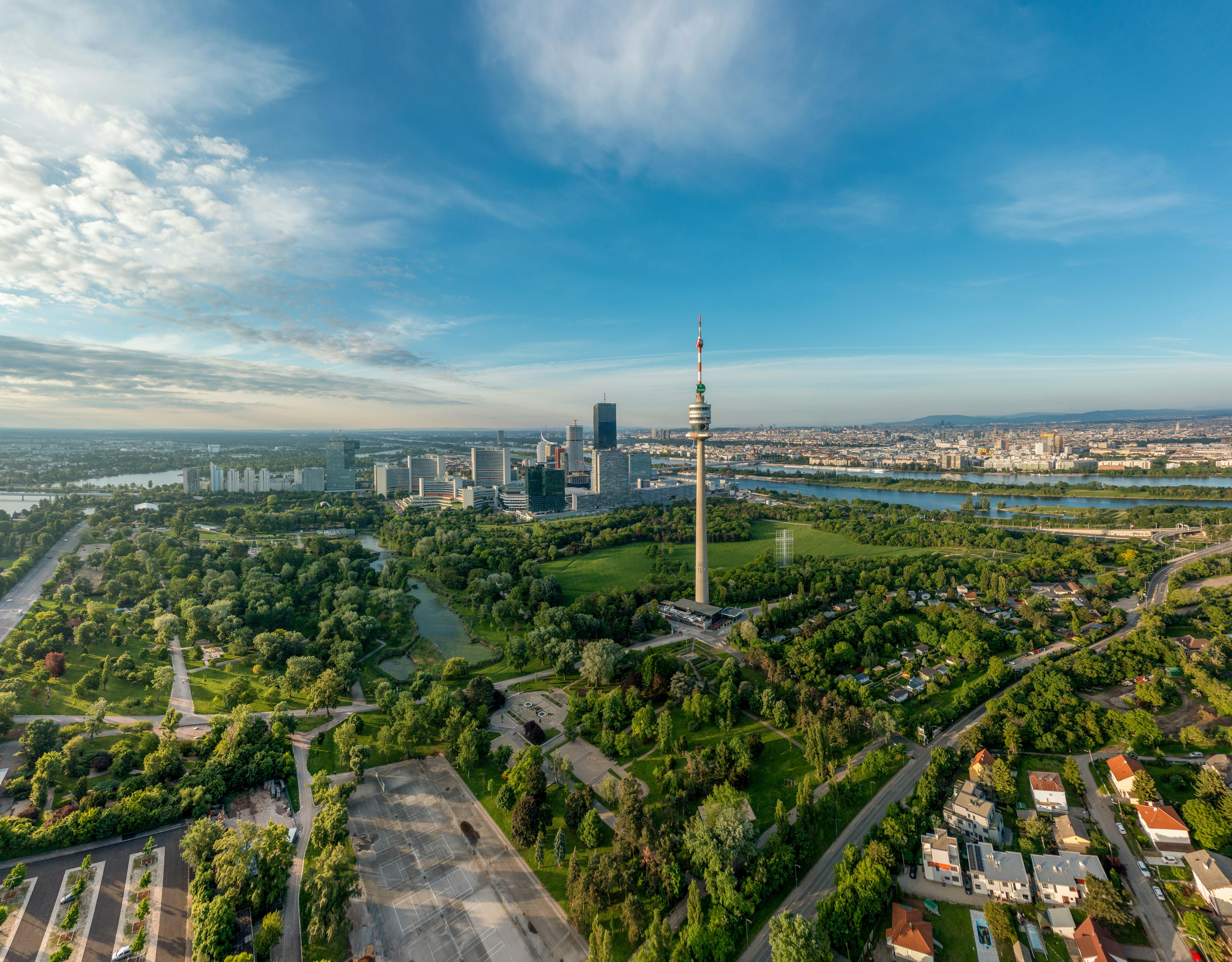 Aerial view of a cityscape with a prominent tower, urban buildings, green parks, and a river under a blue sky.