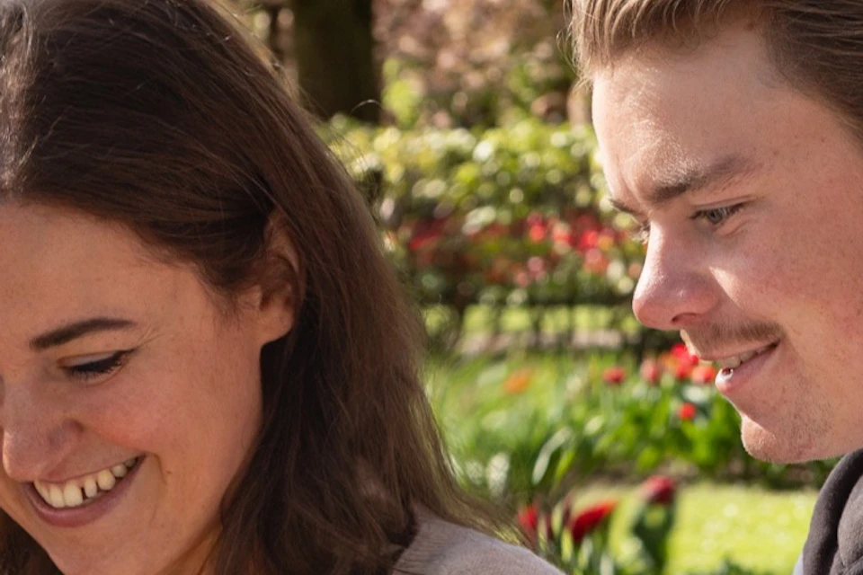 Two people sitting outdoors in a garden, looking at a colorful map together, with flowers and trees in the background.
