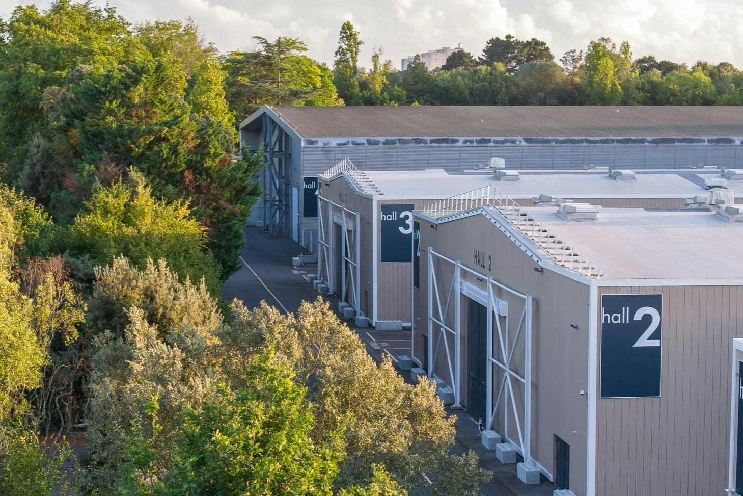 Edifícios industriais denominados «Pavilhão 2» e «Pavilhão 3», rodeados por árvores, sob um céu parcialmente nublado.