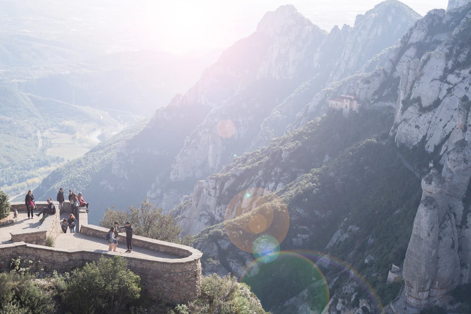 Two people stand on a stone platform overlooking a mountain landscape with steep cliffs and a distant building.