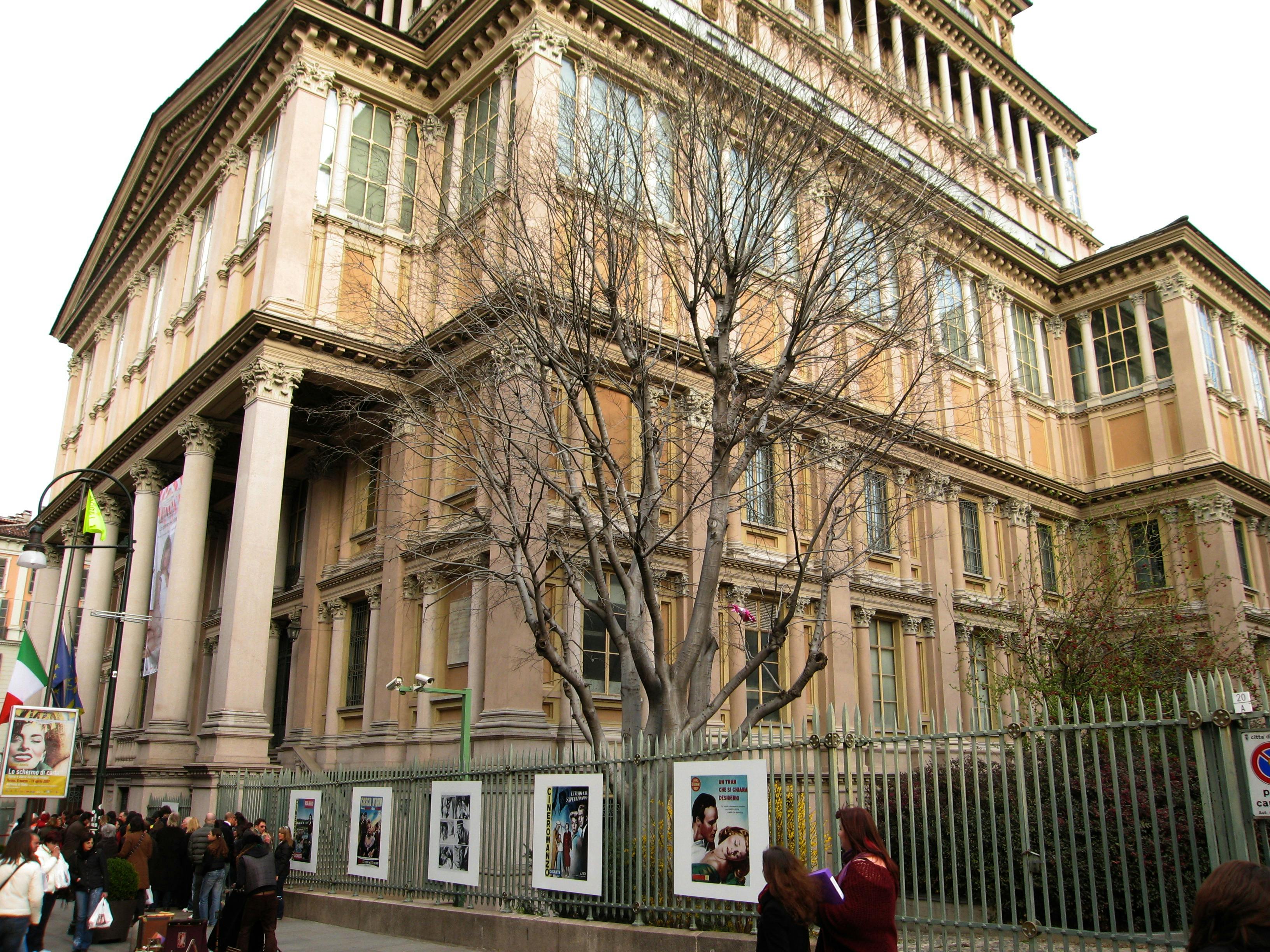 Ein großes, verziertes Gebäude mit hohen Fenstern und einem blattlosen Baum davor. Zwei Personen gehen in der Nähe eines Zauns, an dem Plakate hängen.