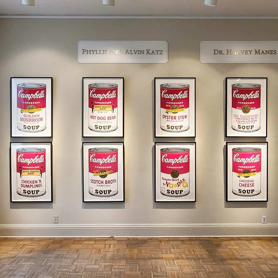 Six framed Campbell's soup can prints hang on a gallery wall with names "Phyllis and Alvin Katz" and "Dr. Harvey Manes" above.