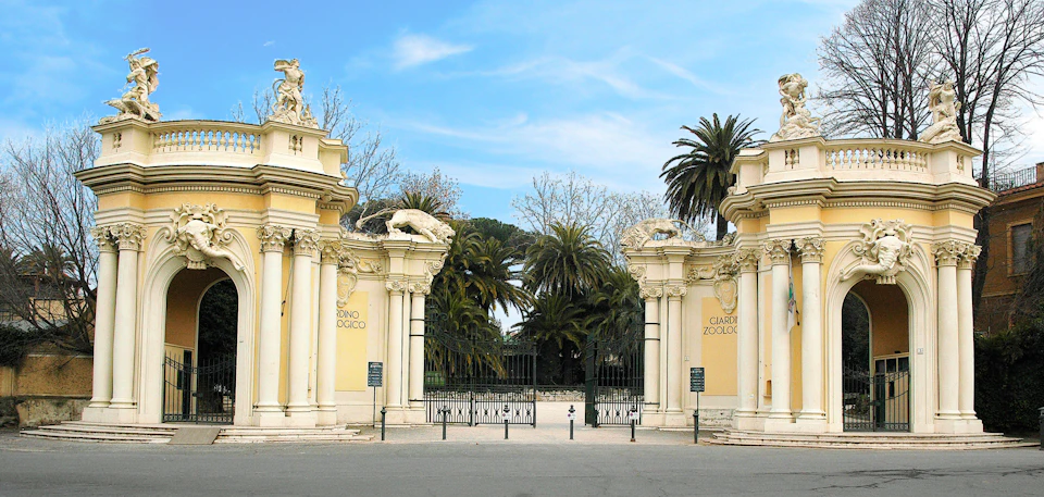 Decorative zoo entrance with ornate columns, statues, iron gates, and "Giardino Zoologico" signs. Palm trees and blue sky visible.
