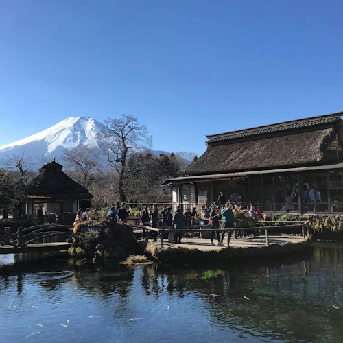 A serene pond with a reflection, wooden buildings with thatched roofs, and Mount Fuji in the background under a clear sky.