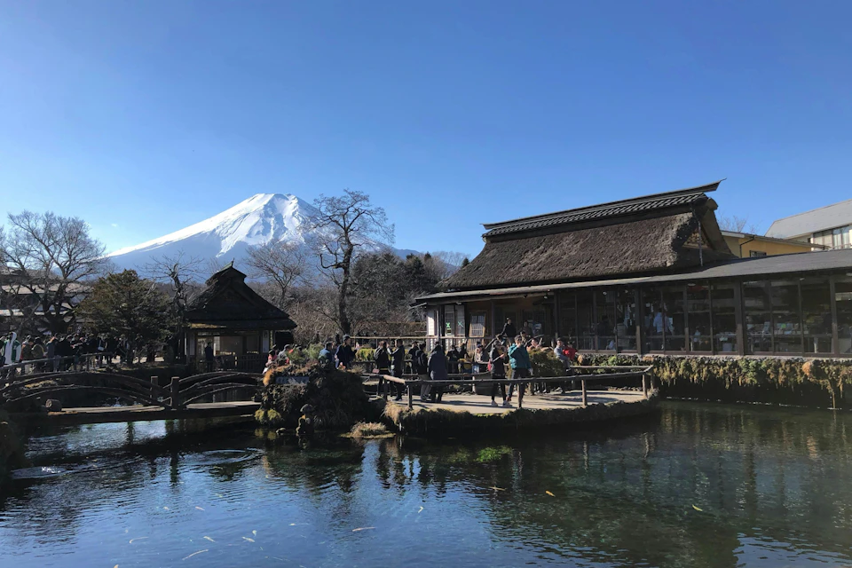 A serene pond with a reflection, wooden buildings with thatched roofs, and Mount Fuji in the background under a clear sky.