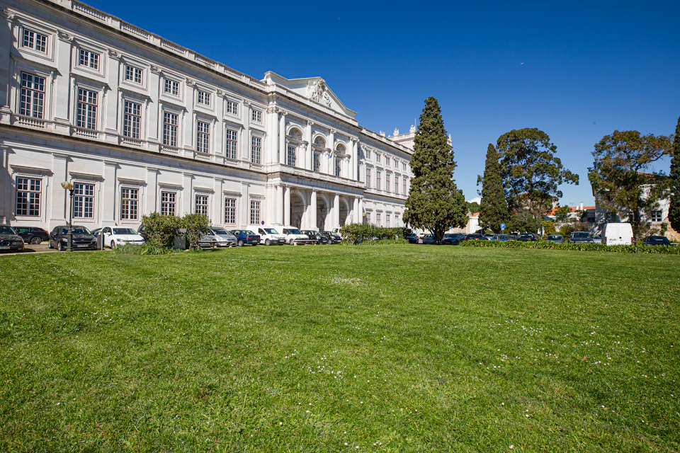 A large, historic building with many windows, a manicured lawn, parked cars, and tall trees under a clear blue sky.