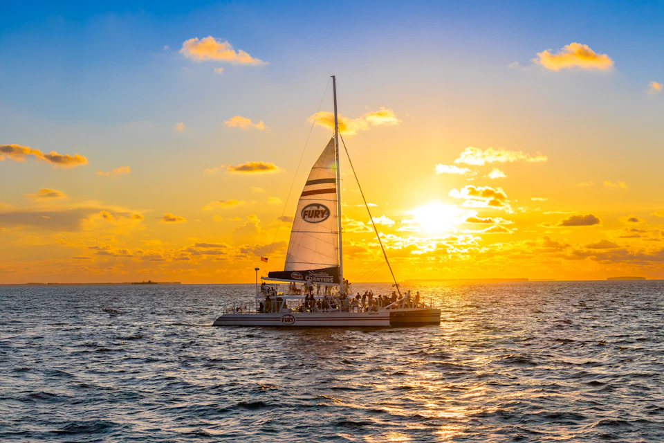 A catamaran with people on board sails on the ocean during sunset, with a large sail and the sun low on the horizon.