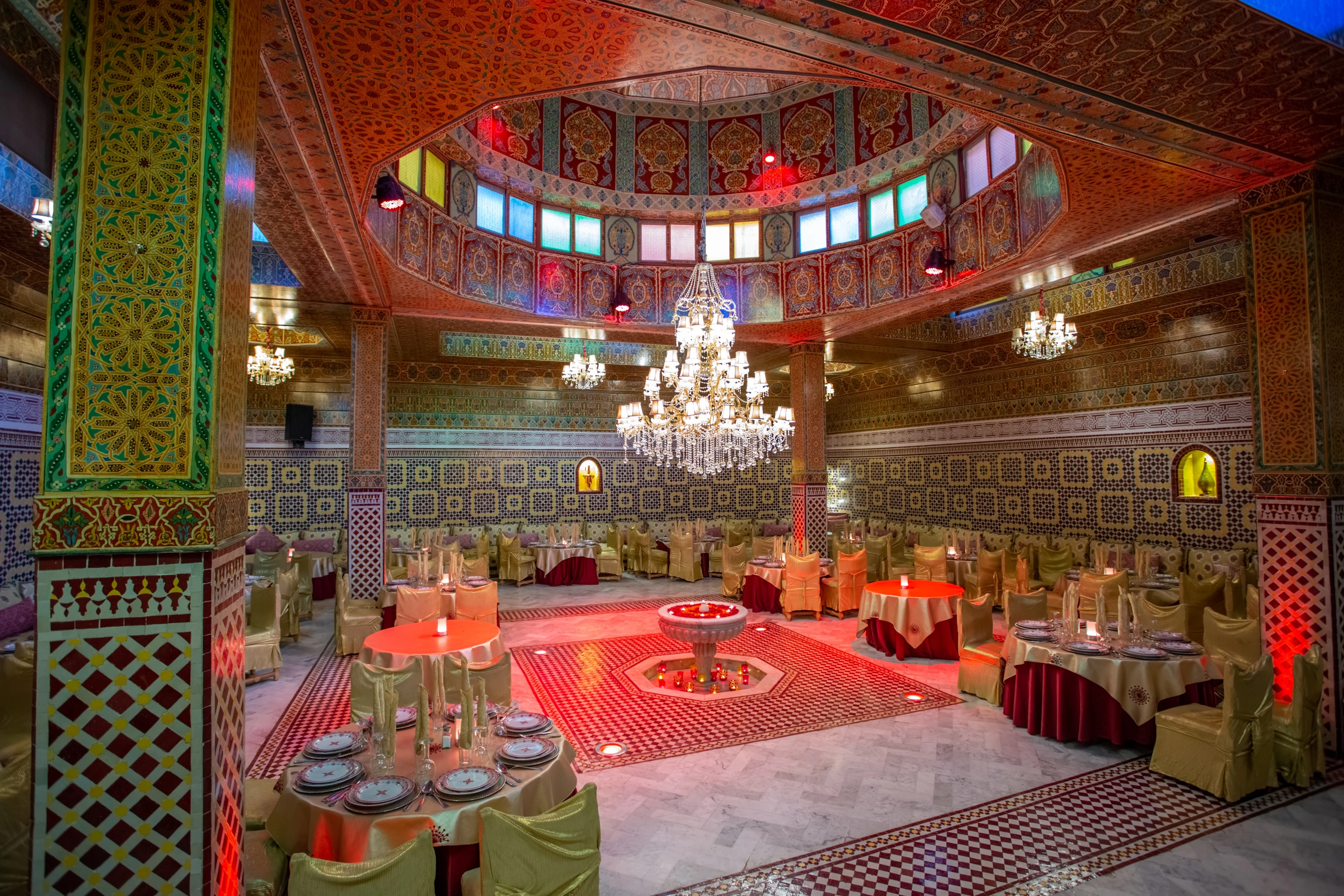 An ornate Moroccan-style dining hall with intricate tilework, colorful stained glass, multiple chandeliers, and round tables set for guests.