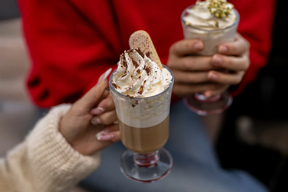 Two people holding glasses of latte topped with whipped cream, a biscotti, and decorative toppings. One person wears a red sweater.
