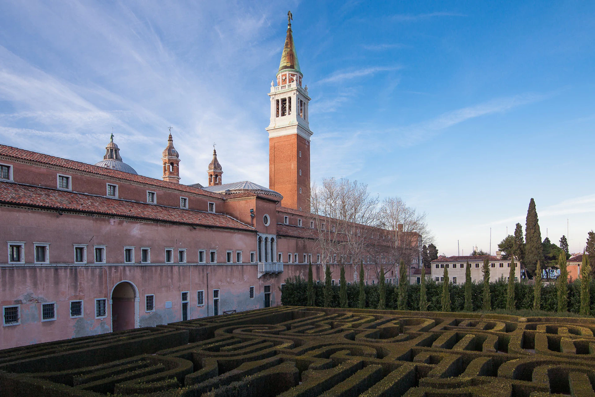 A formal garden maze in front of a large historic building with a tall bell tower under a blue sky.