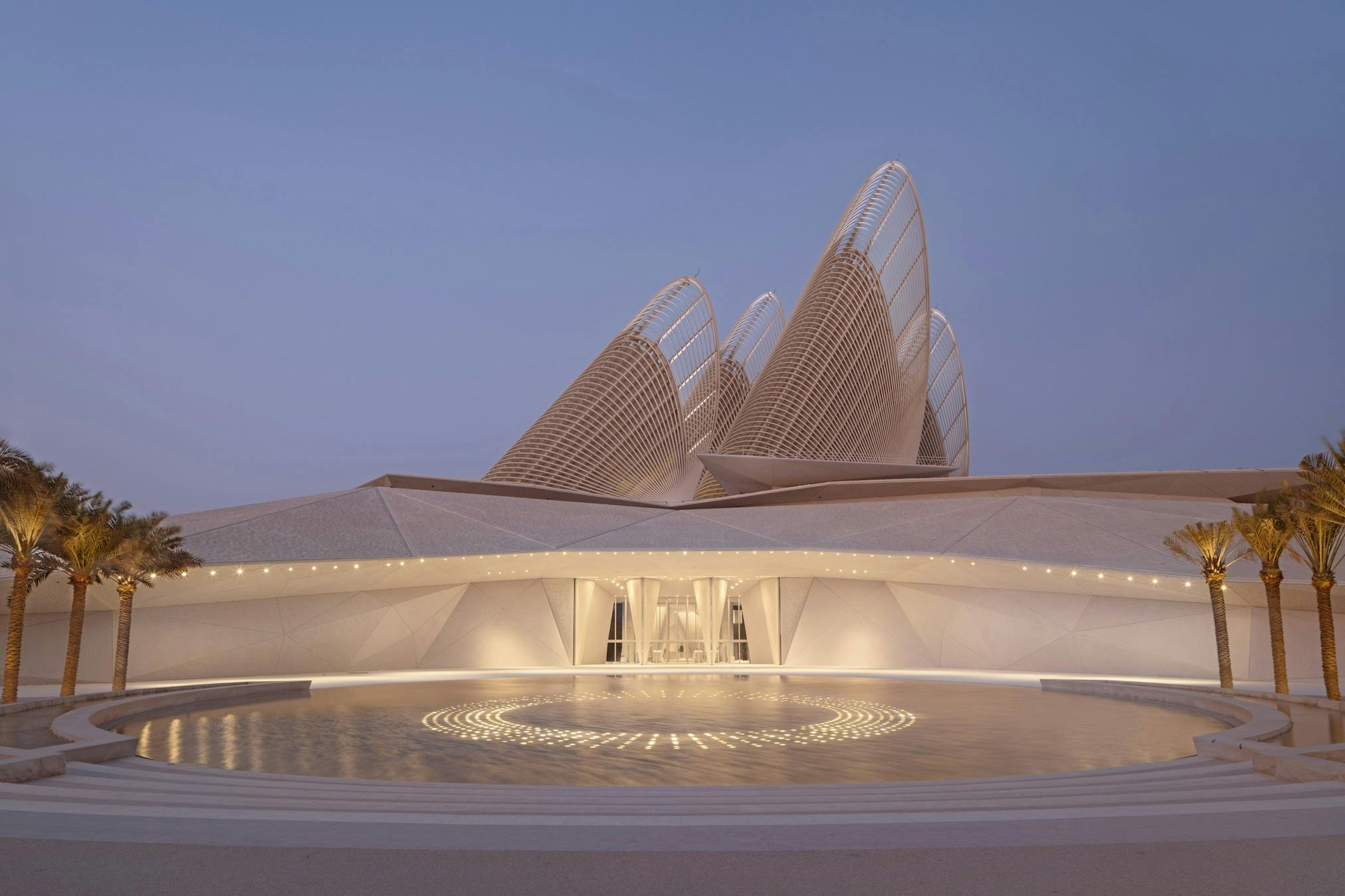 Moderní architektonická budova se zakřivenými mřížkovitými strukturami a kruhovým bazénem s vodou před sebou, pod soumrakem.