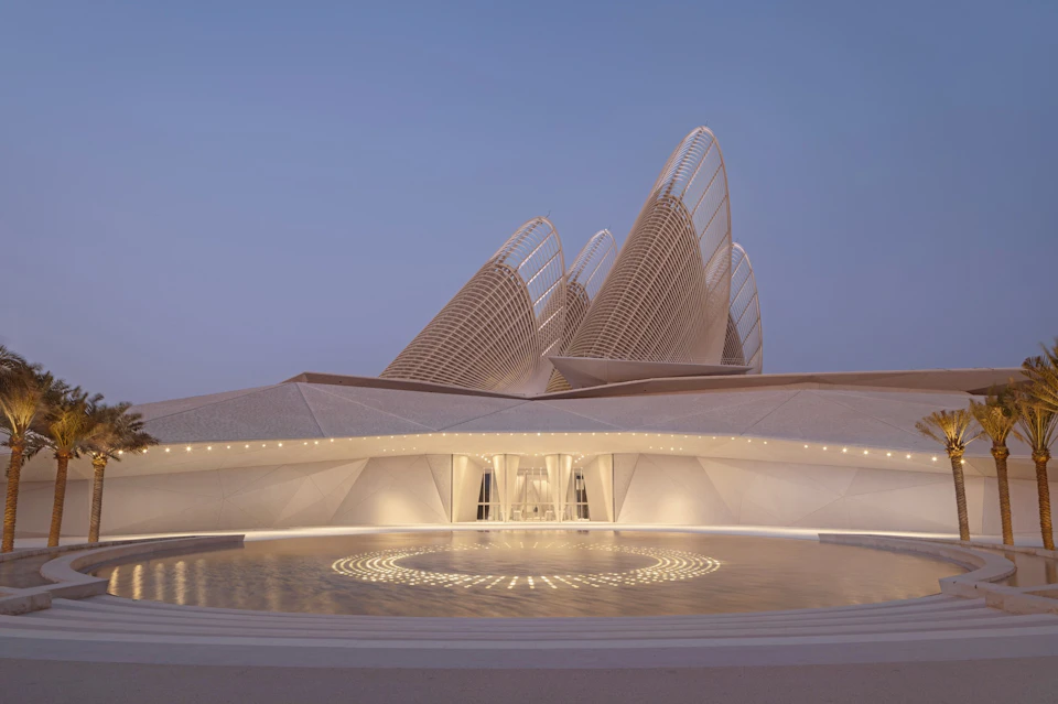 Modern architectural structure with curved, sail-like elements under a twilight sky, featuring a circular lit pattern in front.