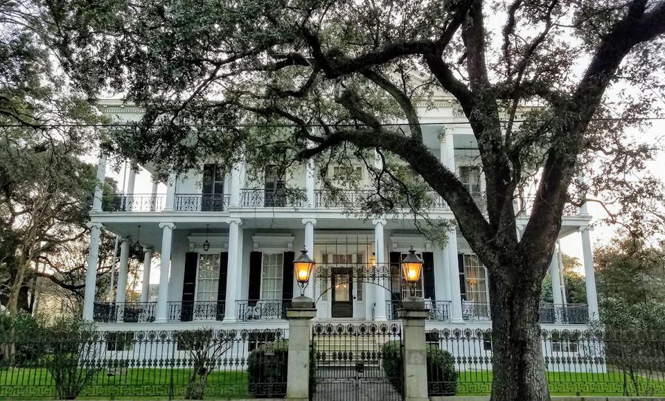 A large, classic two-story white house with columns, iron railings, and a gated entrance, partially framed by tree branches.