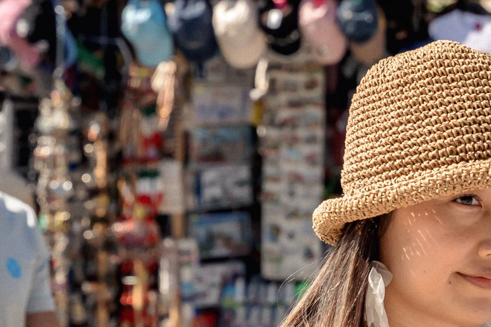 A couple walking near a market with a variety of items on display. Both are wearing summer clothing and hats.