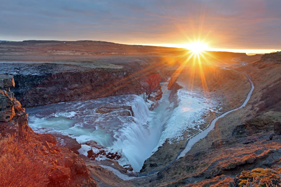A powerful waterfall cascades through a rocky canyon at sunset, with the sun casting a golden glow over the landscape.