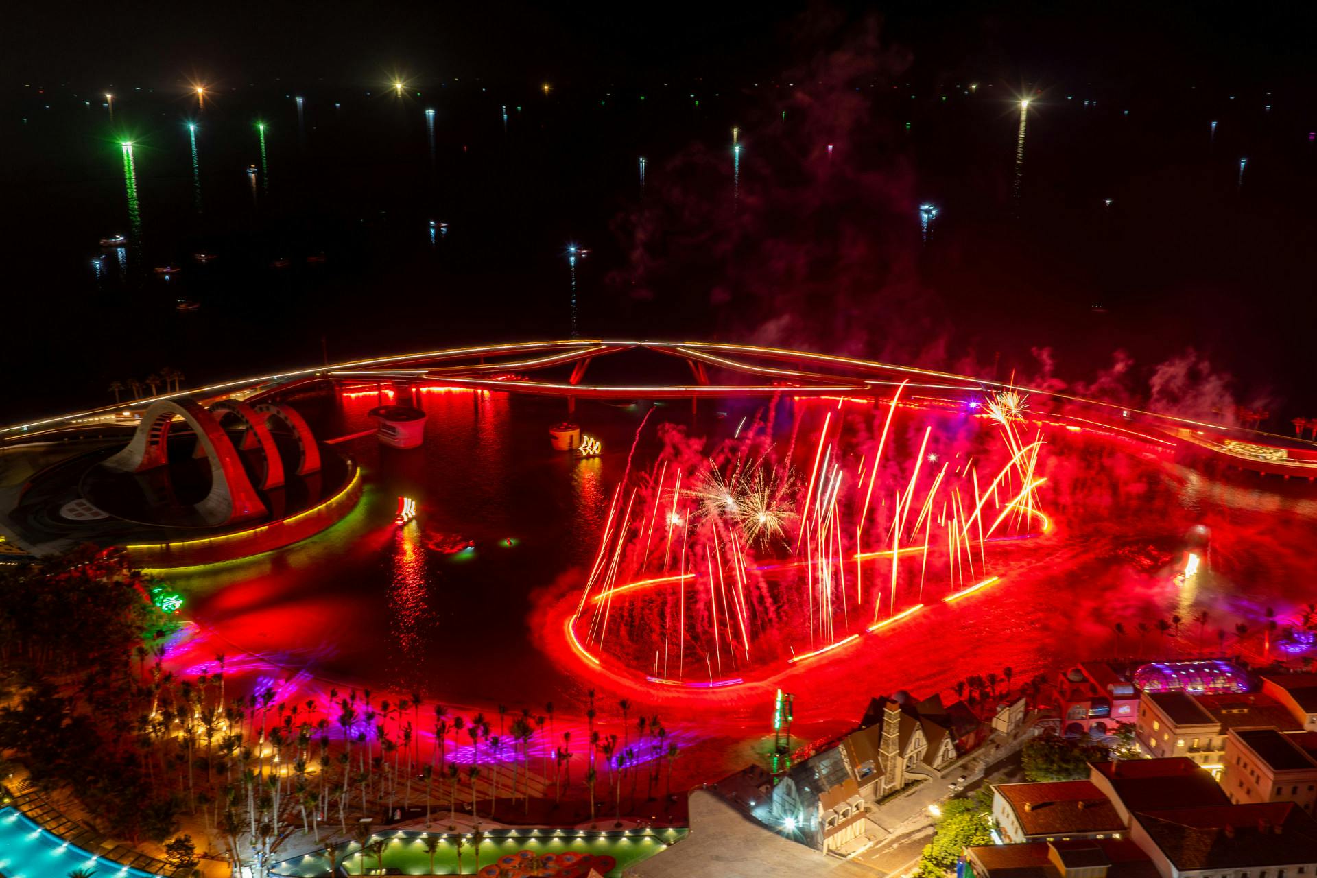 A vibrant nighttime display featuring a bridge lit by red lights, fireworks, and illuminated water features.
