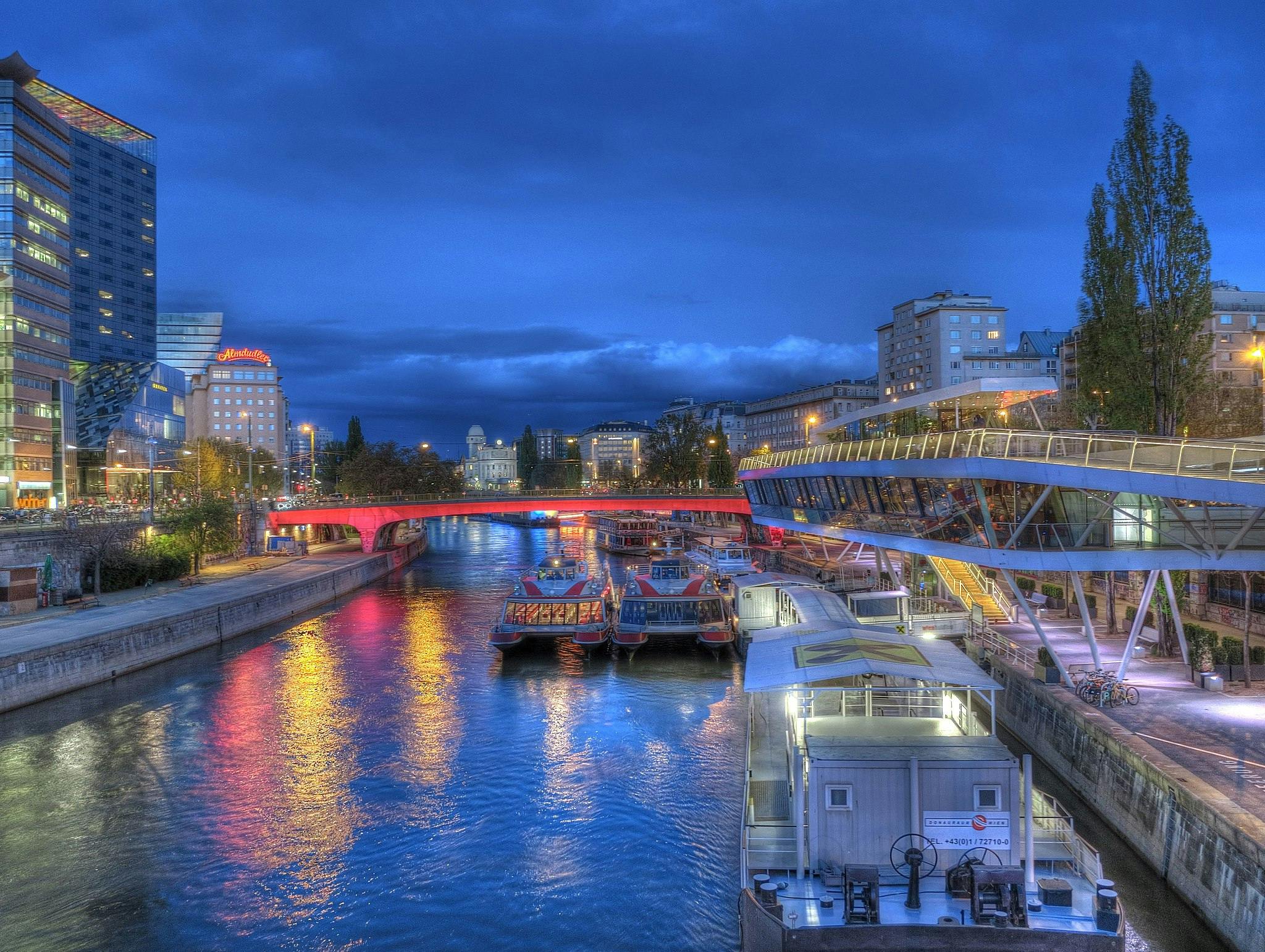 Un canal urbano muy iluminado al atardecer, con barcos iluminados, un puente moderno y edificios altos que se reflejan en el agua.