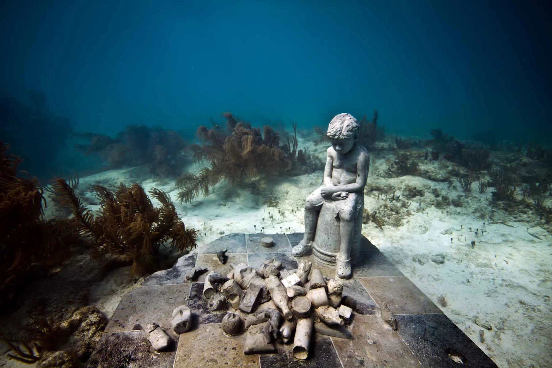 An underwater sculpture of a seated child, surrounded by broken objects on a tiled surface, with aquatic plants in the background.